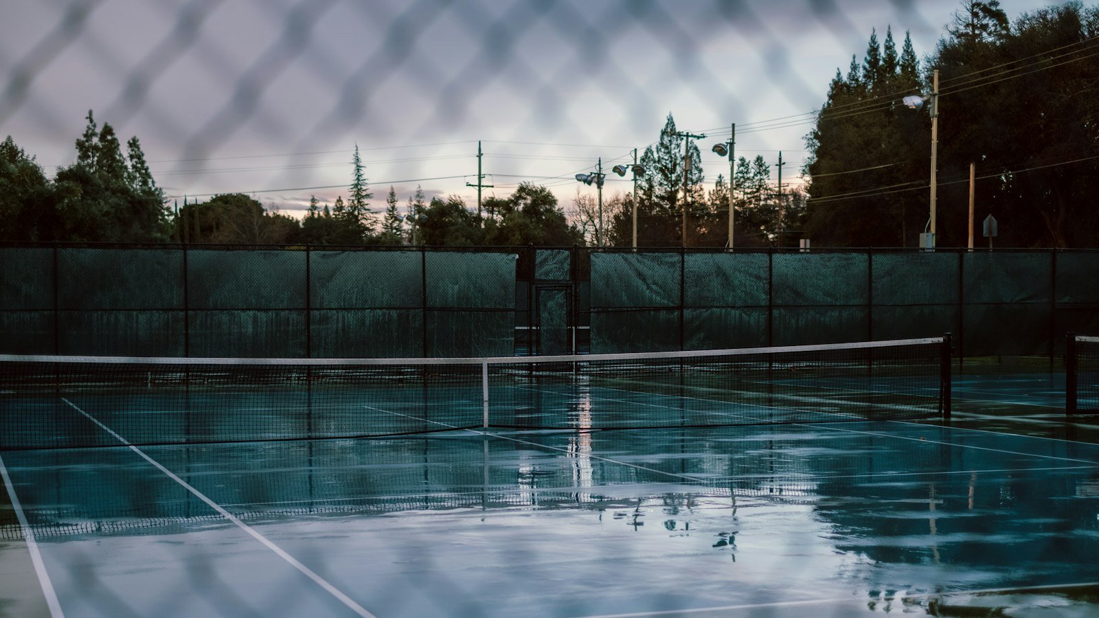 Wet tennis court after rain, seen through the fence