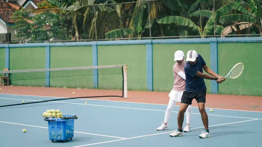 Tennis coach guiding a player's forehand on an outdoor court, with a basket of balls nearby