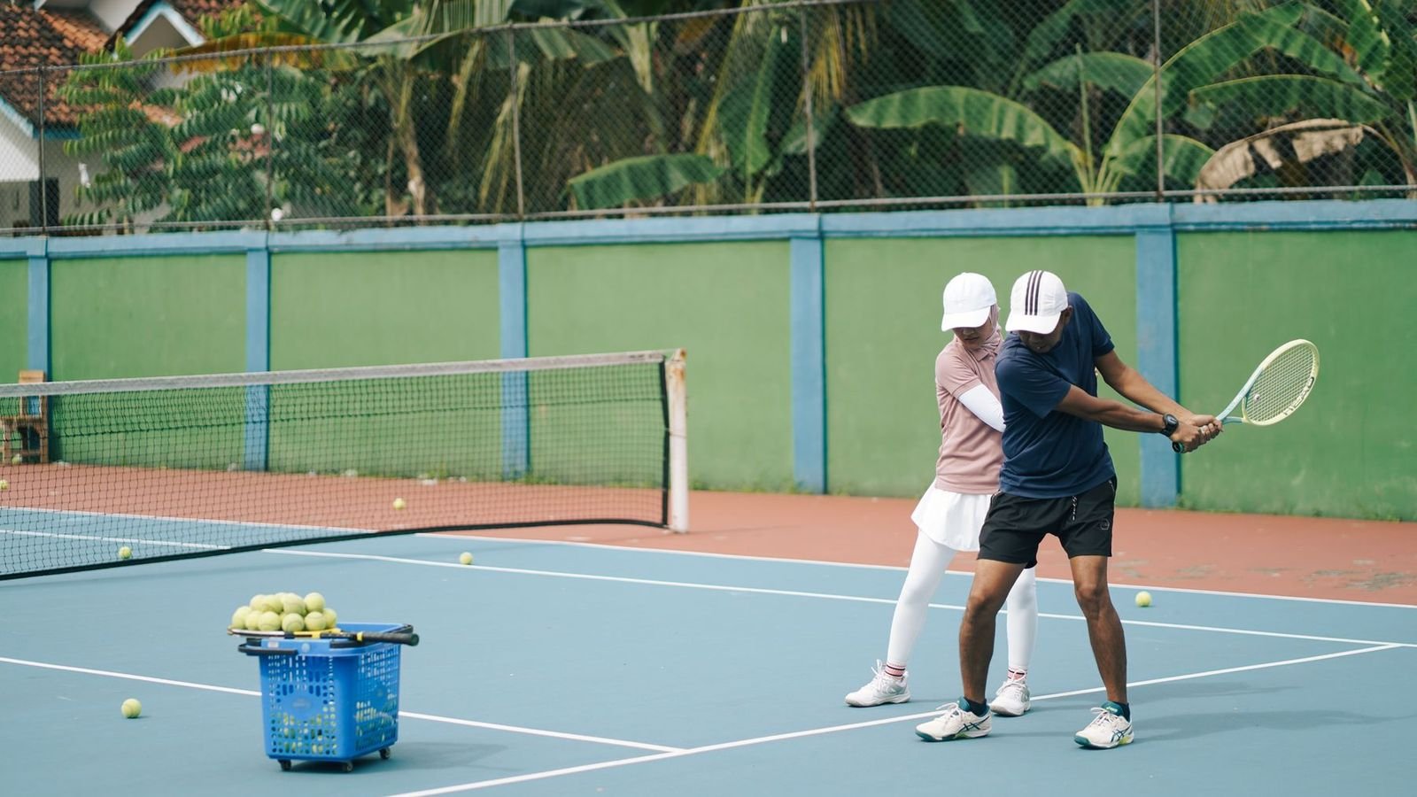 Tennis coach guiding a player's forehand on an outdoor court, with a basket of balls nearby